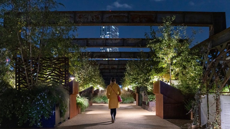 A woman in a yellow coat walking down Castlefield Viaduct in Manchester at night surrounded by lanterns in the gardens.